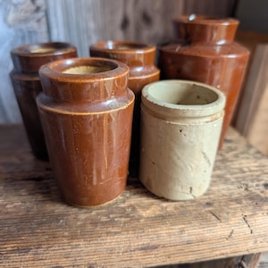 Könnte beinhalten: Eine Sammlung antiker brauner Steinzeuggläser und eine Vase. Die Gläser variieren in Größe und Form, mit einem zylindrischen Design und einem hellbraunen Glas. Die Artikel sind auf einer rustikalen Holzoberfläche angeordnet und zeigen ihr gealtertes Aussehen.