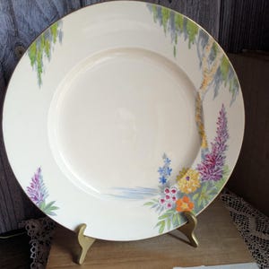 May include: Cream ceramic plate with a gold rim, decorated with a painted landscape. The design includes a tree, flowers in purple, blue, and orange, and green weeping foliage. The plate is displayed on a gold stand, likely for display.