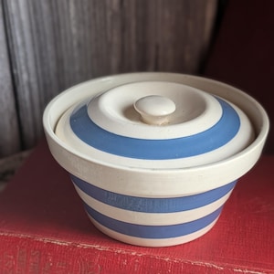 May include: A ceramic bowl with a lid, featuring alternating bands of blue and white. The bowl has a rounded shape with a matching lid and a small knob. The design is reminiscent of vintage kitchenware.