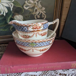 Könnte beinhalten: Ein vintage Creme-farbener Milchkännchen- und Zuckerdose-Set mit einem blauen und rosa Blumenmuster. Das Set verfügt über einen goldenen Rand und ein zartes Blumenmuster.