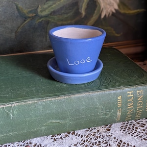 Könnte beinhalten: Ein kleiner, blauer Keramik-Blumentopf mit passender Untertasse. Das Wort "Love" ist handschriftlich auf den Topf geschrieben. Der Topf steht auf einem grünen Buch mit einer weißen Spitzendecke.