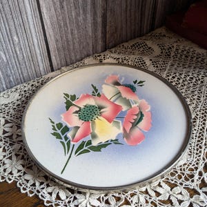Könnte beinhalten: Ein runder, antiker Servierteller mit floralem Design. Der Teller hat einen weißen Hintergrund mit rosa, gelben und grünen Blumen. Die Blumen sind vor einem hellblauen Farbverlauf angeordnet. Der Teller hat einen silberfarbenen Metallrand.