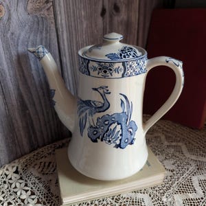 May include: A white ceramic teapot with a blue floral and bird design. The teapot has a curved spout, a handle, and a lid with a matching pattern. The design features a peacock and floral elements. The teapot is sitting on a book.