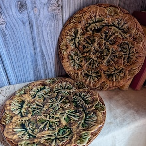 May include: Two round ceramic plates featuring a raised, textured relief pattern of overlapping green and brown leaves. The glaze has a glossy finish with deep green centers and earthy brown edges, creating a dense, botanical design across the surface.