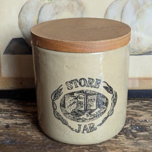 Peut inclure: Un pot de rangement en céramique beige avec un couvercle en bois. Le pot présente un graphisme noir et blanc avec les mots "STORE JAR" et des images de café, de thé et de riz. Le pot cylindrique a une esthétique rustique et vintage.