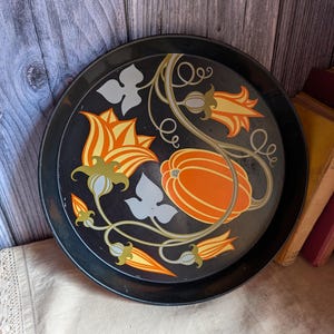 May include: A round black metal serving tray featuring a central graphic design of an orange pumpkin surrounded by stylized orange and yellow flowers with green vines and light gray leaves. The tray has a smooth finish and a raised circular rim.