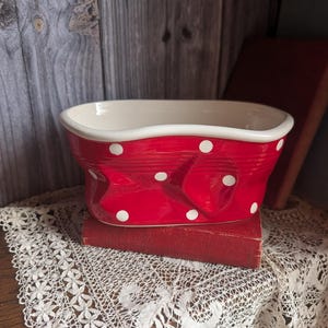 Puede incluir: Un cuenco de cerámica rojo con lunares blancos y un interior blanco. El cuenco tiene un diseño en forma de lazo en la parte delantera y está sobre un libro rojo. El cuenco está sobre un tapete de encaje.