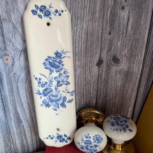 Op de afbeelding: Een verticale rechthoekige keramische deurplaat en twee bijpassende ronde deurknoppen, allemaal met een crèmekleurige basis en een blauw bloemmotief. De deurknoppen hebben gepolijste goudkleurige metalen voetstukken en onderdelen.