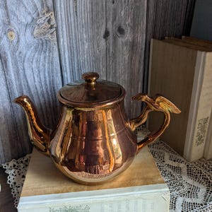 Puede incluir: Una tetera de color cobre con tapa y asa decorativa. La tetera está sobre un libro. La tetera tiene una superficie brillante y reflectante. El asa tiene un diseño en forma de pájaro.