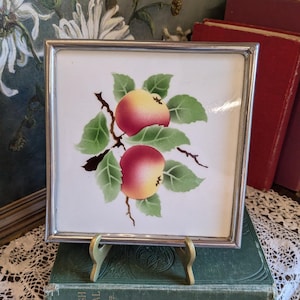 Könnte beinhalten: Quadratische Keramikfliese mit silbernem Metallrahmen. Die Fliese zeigt eine Malerei von zwei rot-gelben Äpfeln mit grünen Blättern auf einem Zweig. Die Fliese wird auf einem kleinen goldfarbenen Ständer präsentiert, der einen Vintage-Look hat.