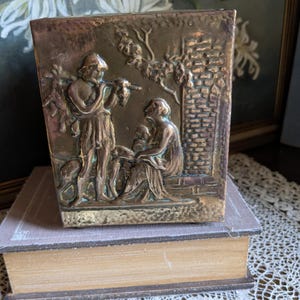 Könnte beinhalten: Ein antiker Messing-Pflanztopf mit einem geprägten Design von zwei Figuren in einer Gartenlandschaft. Der Topf hat eine rechteckige Form und eine strukturierte Oberfläche.