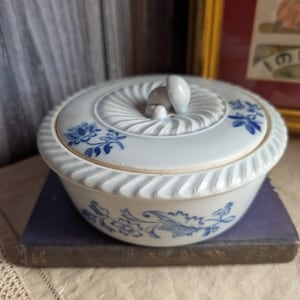Peut inclure: Un plat de service en céramique blanche avec un couvercle, orné de motifs floraux bleus. Le plat a un bord cannelé et un bouton sur le couvercle. Il est posé sur un livre ancien, évoquant un style classique.