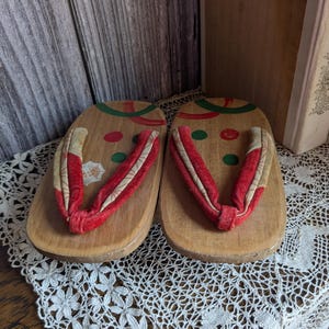 May include: A pair of wooden geta sandals with red and cream fabric straps. The wooden platforms are decorated with green and red painted accents. The sandals are displayed on a white lace doily.
