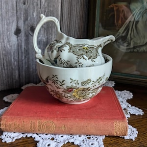 Könnte beinhalten: Ein vintage Creme-farbener Sahnegießer und Zuckerdose Set mit einem floralen Muster in Braun-, Grün- und Gelbtönen. Das Set steht auf einem roten Buch mit einem goldenen Design.