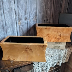 Könnte beinhalten: Zwei rechteckige Holzkisten mit hellbraunem Maserholz-Finish und schwarzem Innenraum. Die Kisten werden auf einer dunklen Holzoberfläche präsentiert, mit einem dekorativen Spitzentuch und Büchern.