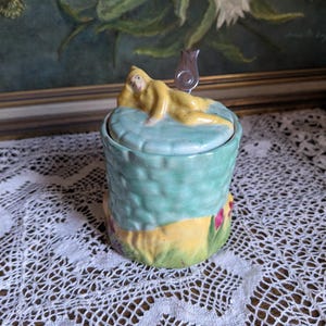 Peut inclure: Un pot en céramique avec un motif floral vert et jaune. Le couvercle présente une figure sculptée d'une femme allongée avec une cuillère en argent.