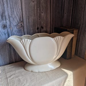 Può includere: Vaso vintage in ceramica color crema, con bordo smerlato e base a piedistallo. Il vaso ha un design simmetrico con nervature verticali e una sezione centrale arrotondata. È appoggiato su una tovaglia chiara su sfondo in legno.
