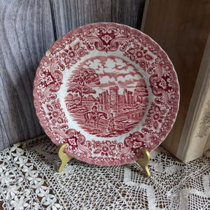 May include: A red and white decorative plate with a detailed landscape scene featuring a castle, trees, and figures on horseback. The plate has a scalloped edge with floral and scrollwork patterns, and is displayed on a gold-coloured stand.