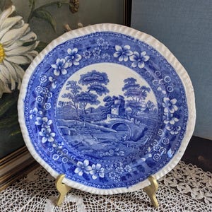 Op de afbeelding: Blauw en wit decoratief bord met een landschap met een brug, bomen en een kasteel. Het bord heeft een bloemenrand en een geschulpte rand. Het wordt gepresenteerd op een goudkleurige standaard.