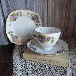 Può includere: Servizio da tè bianco con tazza, piattino e piatto con design floreale viola e bordi dorati. La tazza è su un piattino e il piatto a sinistra. Il set è su un libro vintage.