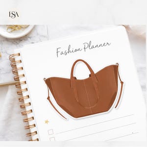 May include: A spiral-bound planner with the words "Fashion Planner" in cursive. A brown handbag illustration is centered on the page. The planner has a gold spiral binding and a white cover with a minimalist design.