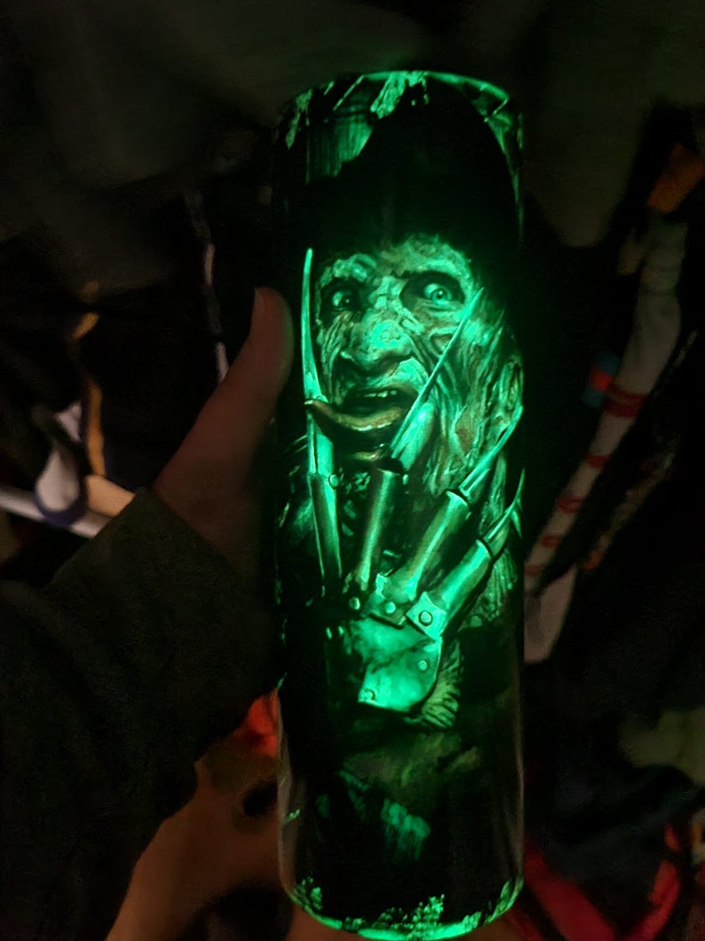Puede incluir: Un vaso verde que brilla en la oscuridad con un gr&aacute;fico de Freddy Krueger de la pel&iacute;cula "Pesadilla en Elm Street".