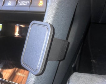 May include: A black and gray car phone mount installed on the dashboard. The mount has a rectangular pad with a dark gray border and a black bracket. The dashboard is gray with various buttons and controls visible.