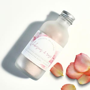 May include: A frosted glass bottle with a silver cap, labeled "Callista Hibiscus & Rose" with floral accents. The bottle contains a pinkish powder and is accompanied by scattered rose petals. The label also reads "Cleansing Grains/Facial Mask with Freshwater Pearl + Vitamin C."