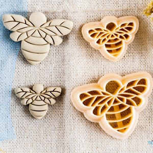 May include: Four beige bee-shaped cookie cutters of varying sizes on a beige linen background. The largest cutter has a detailed design with intricate lines and a honeycomb pattern. The smallest cutter is a simple bee shape with a few lines.