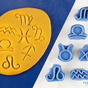 May include: A collection of blue rubber stamps featuring zodiac symbols, including Libra, Taurus, and Sagittarius. A yellow clay-like surface displays embossed zodiac signs. The image is set against a blue and white background.