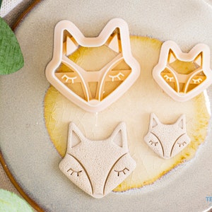 Può includere: Tre stampini per biscotti a forma di volpe su un piatto beige. Lo stampino più grande è al centro, con due stampini più piccoli su entrambi i lati. Tutti e tre gli stampini sono in plastica marrone chiaro.