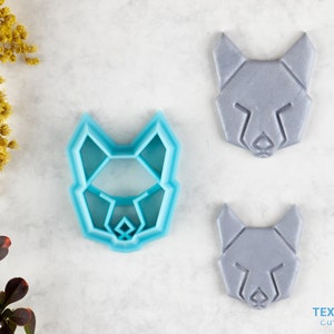 Puede incluir: Un cortador de galletas de plástico azul claro con forma de cabeza de lobo geométrica, junto a dos galletas grises con forma de cabeza de lobo. El cortador y las galletas están sobre una superficie blanca con follaje amarillo y rojo.