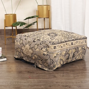 May include: A gray and beige patterned ottoman with a zipper closure. The ottoman is sitting on a wooden floor.