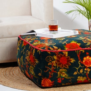 May include: A dark green, floral patterned, square ottoman with a red trim. The ottoman is sitting on a woven rug.