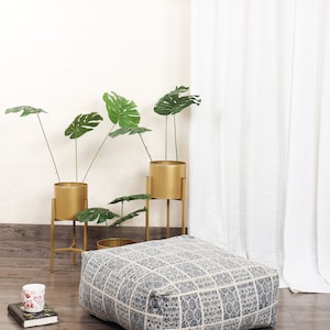 May include: A blue and white patterned floor cushion with a square shape. The cushion is sitting on a wooden floor in front of a white wall and a white curtain. There are three potted plants with green leaves in gold metal stands behind the cushion.