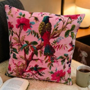 May include: Pink square throw pillow with a colorful bird and floral pattern. The bird has a green head, red body, and blue tail. The flowers are red and pink, with green leaves. The pillow is on a white surface.