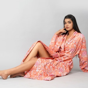 Op de afbeelding: Een vrouw draagt een lange, perzikkleurige badjas met een bloemenprint. De badjas heeft een strikceintuur en lange mouwen. De vrouw zit op een witte achtergrond.