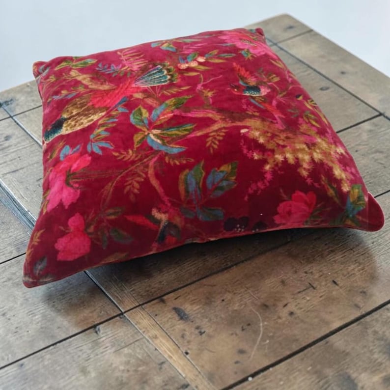 May include: A square, red velvet pillow with a floral and bird pattern. The design features colorful birds, flowers, and foliage in shades of pink, blue, and gold. The pillow is placed on a wooden surface.