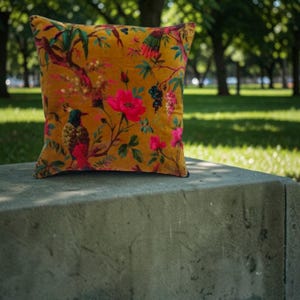 May include: Mustard-colored square throw pillow with a floral and bird pattern. The design features pink flowers, green leaves, and colorful birds on a velvet-like fabric. The pillow is resting on a concrete surface.