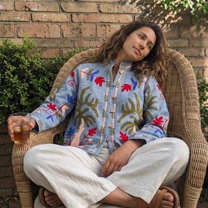 Könnte beinhalten: Eine hellblaue Langarmjacke mit lebendigen Blumen- und Blattstickereien in Rot, Grün und Blau. Die Jacke hat einen Stehkragen, eine Knopfleiste auf der Vorderseite mit kleinen hellen Knöpfen und wird mit einer locker sitzenden weißen Hose kombiniert.