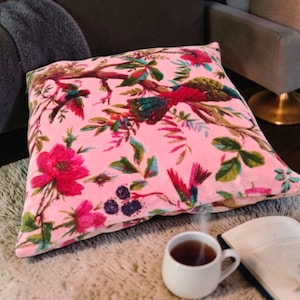 May include: A square pink decorative pillow with a floral and bird pattern. The design features red flowers, green leaves, and colorful birds on branches. The pillow is on a fluffy rug, with a cup of tea and a book in the foreground.