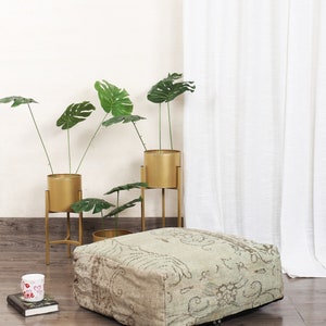 May include: A square floor pouf featuring a faded, light green and beige vintage-style pattern with intricate scrollwork and floral motifs. The pouf has a low-profile, boxy shape with visible stitching along the edges and a small metal zipper pull.