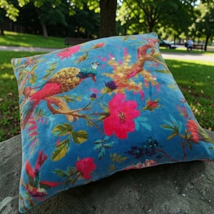 May include: Square decorative pillow with a vibrant floral and bird pattern. The pillow features a teal background with a colorful design of pink flowers, green leaves, and a bird with red, gold, and blue feathers. The pillow is made of a soft, velvety material.