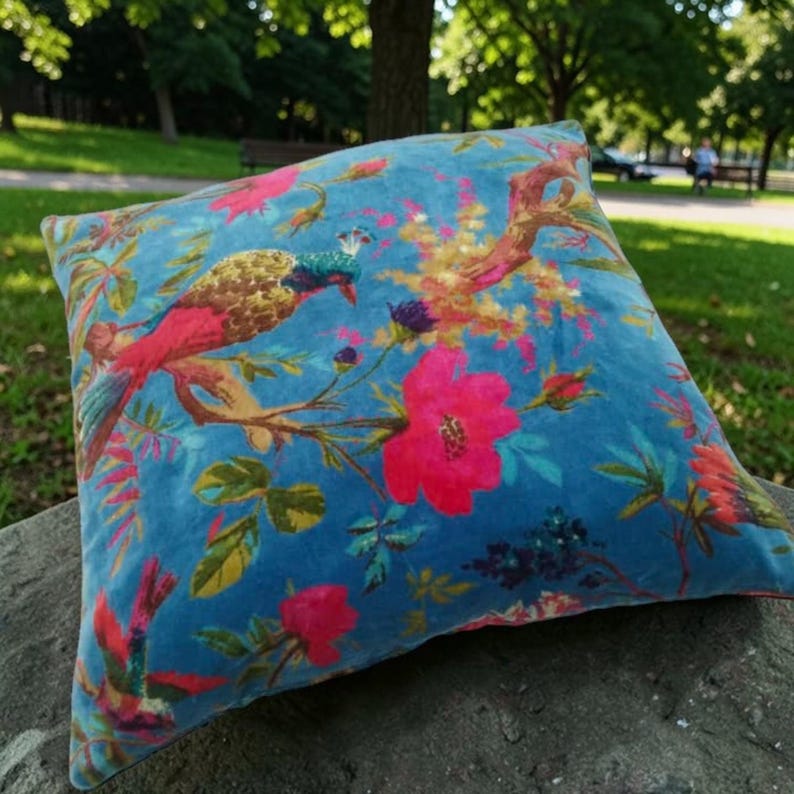 May include: Square decorative pillow with a vibrant floral and bird pattern. The pillow features a teal background with a colorful design of pink flowers, green leaves, and a bird with red, gold, and blue feathers. The pillow is made of a soft, velvety material.