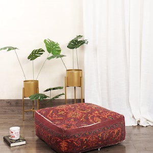 May include: A red and gold patterned floor cushion with a white border. The cushion is sitting on a wooden floor in front of a white curtain and gold plant stands with green plants.
