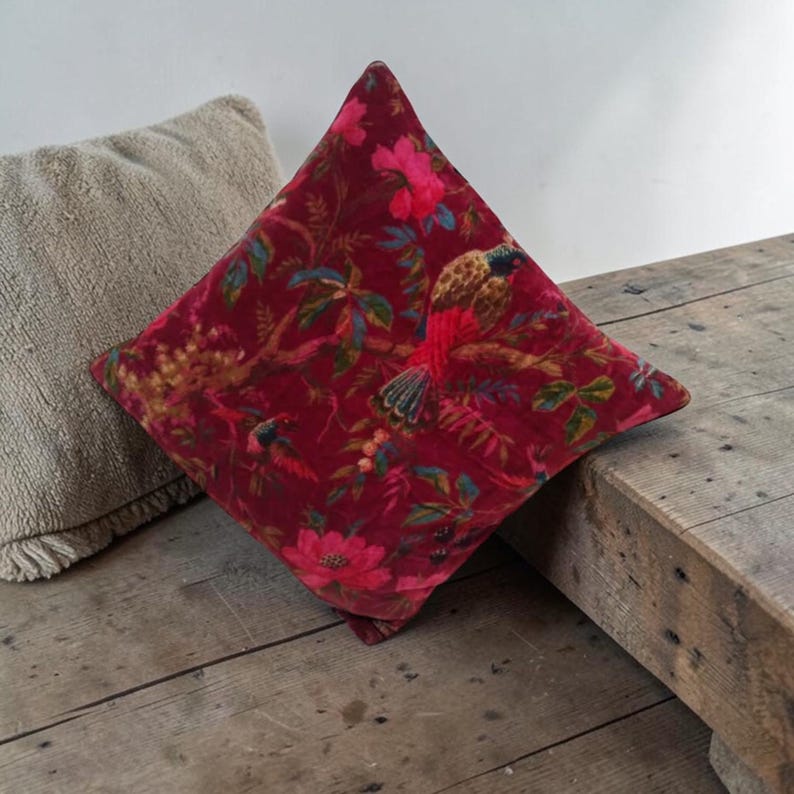 May include: A square, red velvet pillow with a vibrant floral and bird pattern. The design features pink flowers, green leaves, and colorful birds against a deep red background. The pillow is placed on a rustic wooden surface.