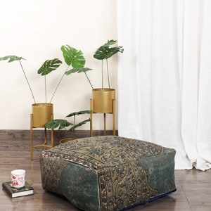 May include: A square, patterned ottoman in shades of green, brown, and blue sits on a wooden floor. The ottoman has a vintage rug design. In the background are gold plant stands with green leafy plants.