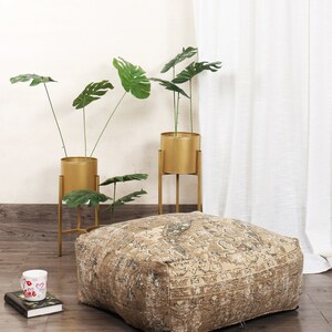 May include: A large, square, beige and brown patterned floor cushion with a decorative, intricate design. The cushion is sitting on a wooden floor in front of two gold metal plant stands with green leafy plants.
