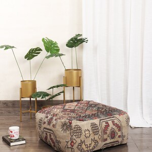 May include: A square, patterned ottoman with a beige, brown, red, and black geometric design. The ottoman is sitting on a wood floor in front of a white wall and a white curtain.