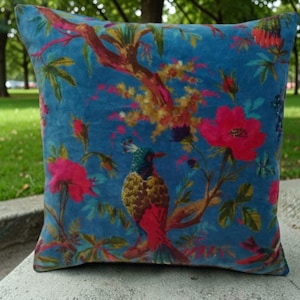May include: A square, blue velvet pillow with a vibrant floral and bird design. The pattern features a colorful bird perched on a branch with pink flowers and green leaves. The pillow is placed on a stone surface, with a blurred green background.